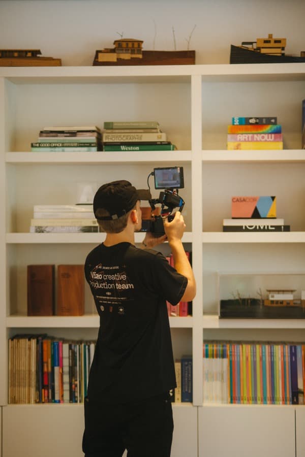 Operador de câmera com camiseta da Vízo filmando com uma câmera em gimbal, diante de uma estante com maquetes de arquitetura e livros de design.