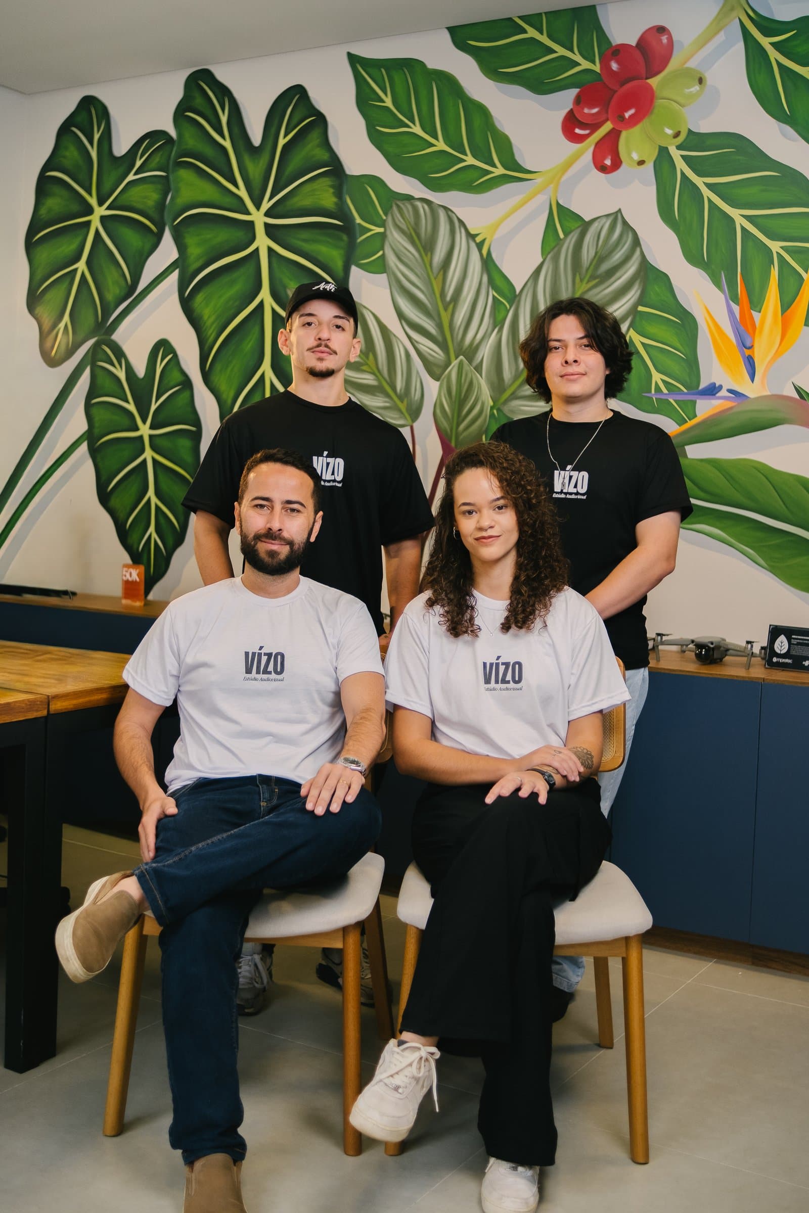 Equipe da Vízo posando diante de um mural de folhagens: duas pessoas em pé atrás e duas sentadas na frente, todas com camisetas da Vízo.